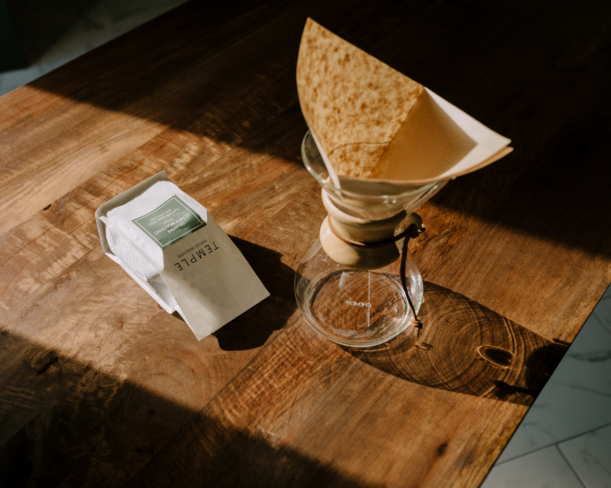 Bag of coffee next to a Chemex brewer with a brown filter on a wooden table.