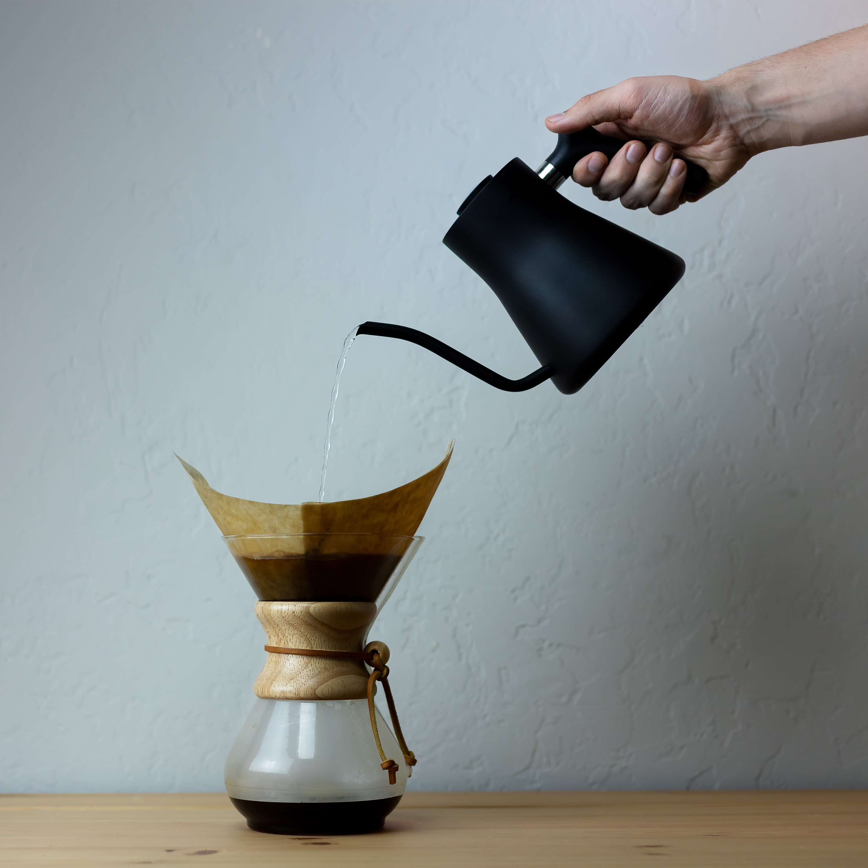 Hand pouring water from a black kettle into a chemex coffee dripper.
