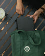 A dark green canvas tote bag with a white circular logo sits on a dark gray slatted table, with a person's hands holding a black notebook inside the bag.