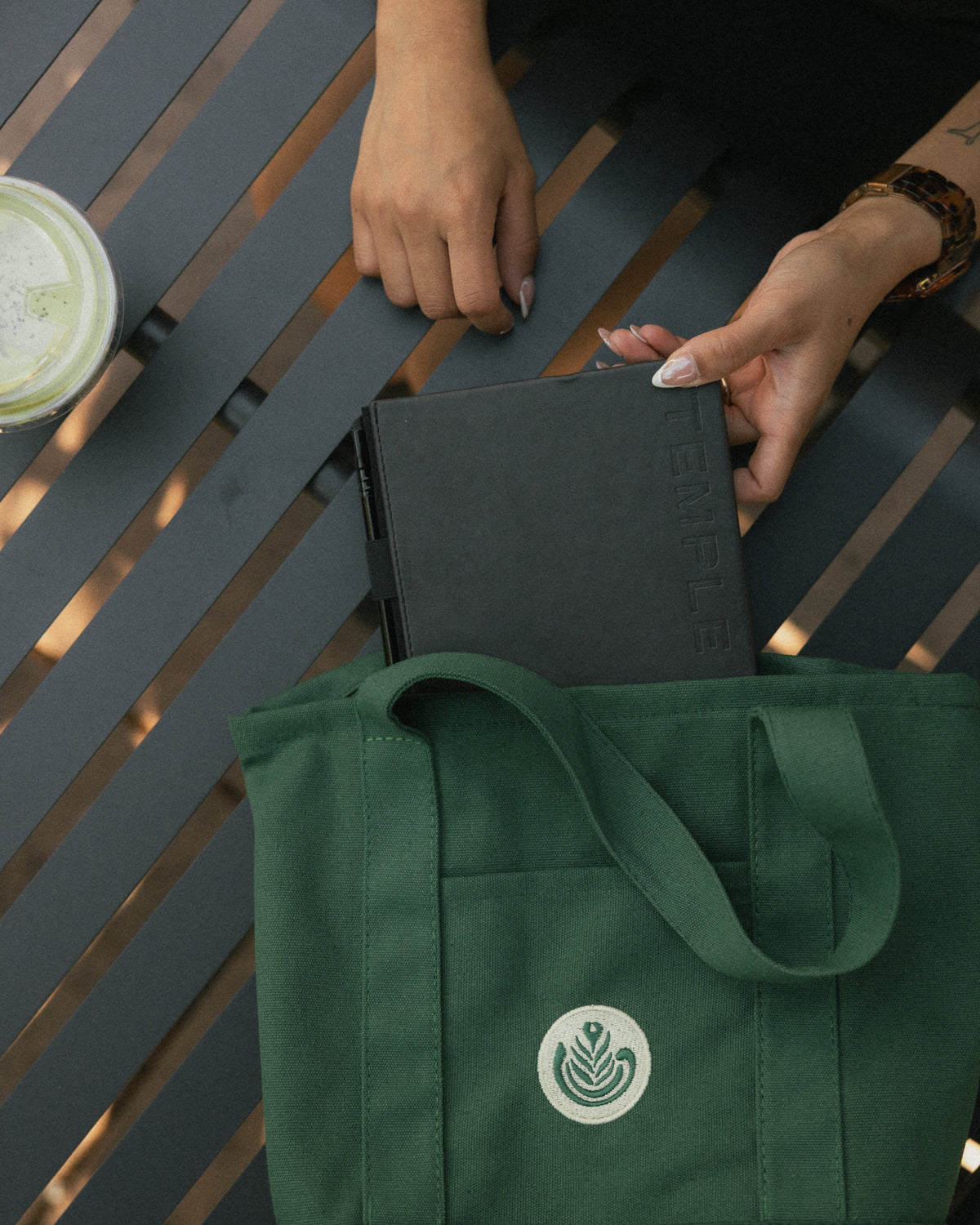 A dark green canvas tote bag with a white circular logo sits on a dark gray slatted table, with a person's hands holding a black notebook inside the bag.
