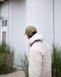 Person wearing a green cap and white jacket standing in front of a white building with greenery.