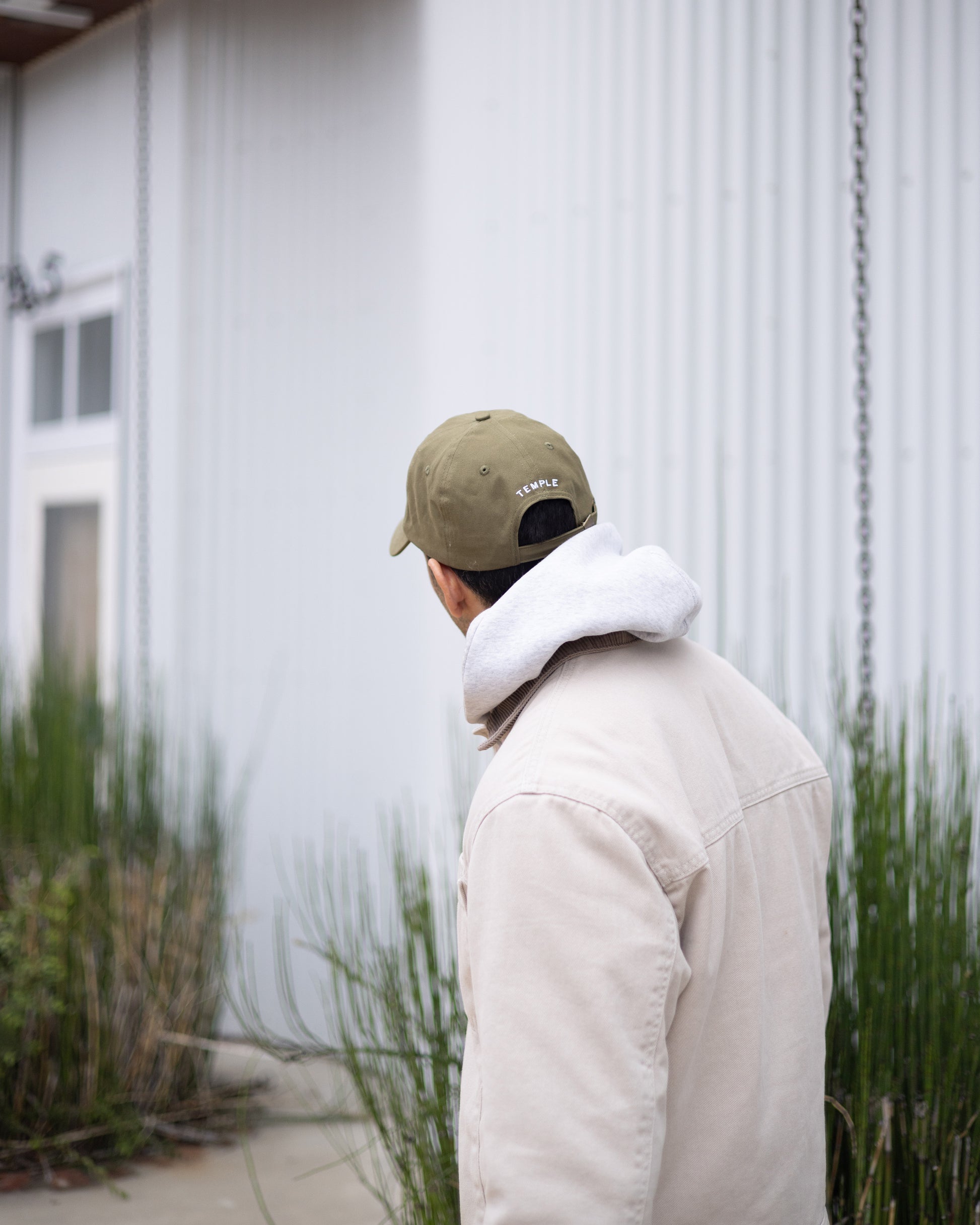 Person wearing a green cap and white jacket standing in front of a white building with greenery.