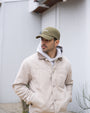 Man wearing a beige jacket and green cap with a logo, standing against a light-colored wall.