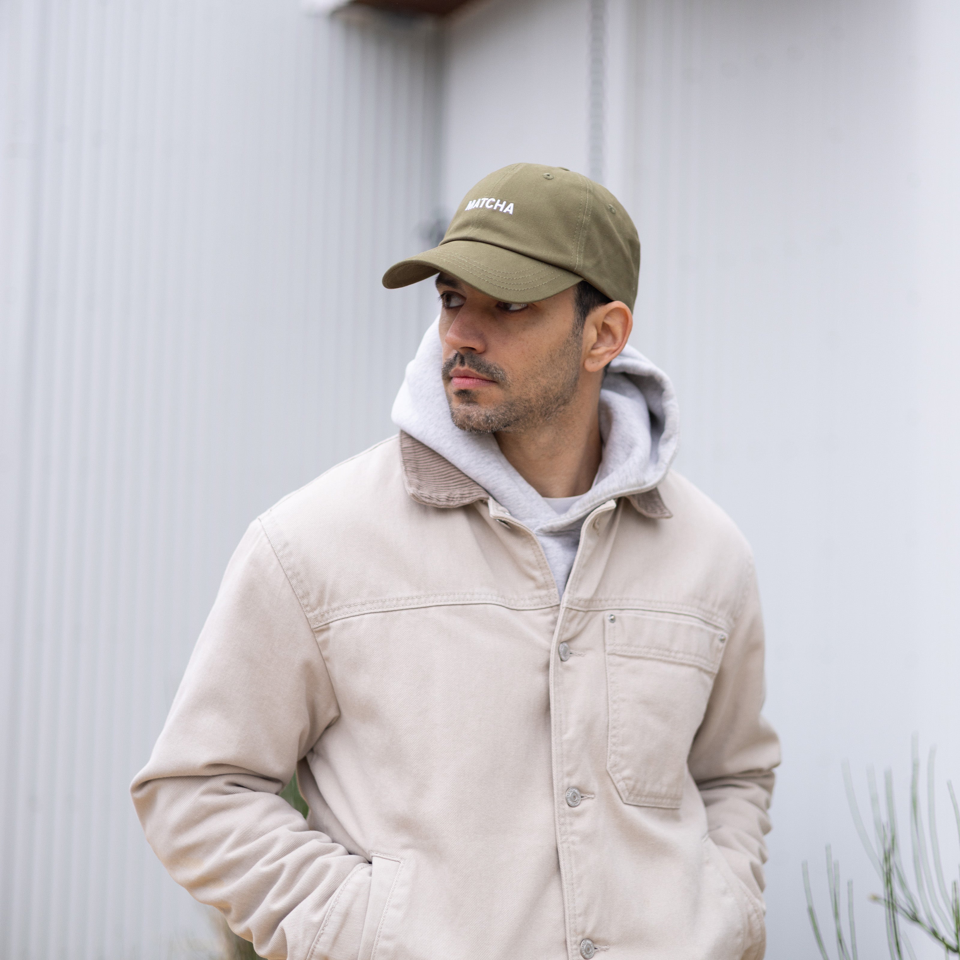 Man wearing a beige jacket and green cap with a logo, standing against a light-colored wall.