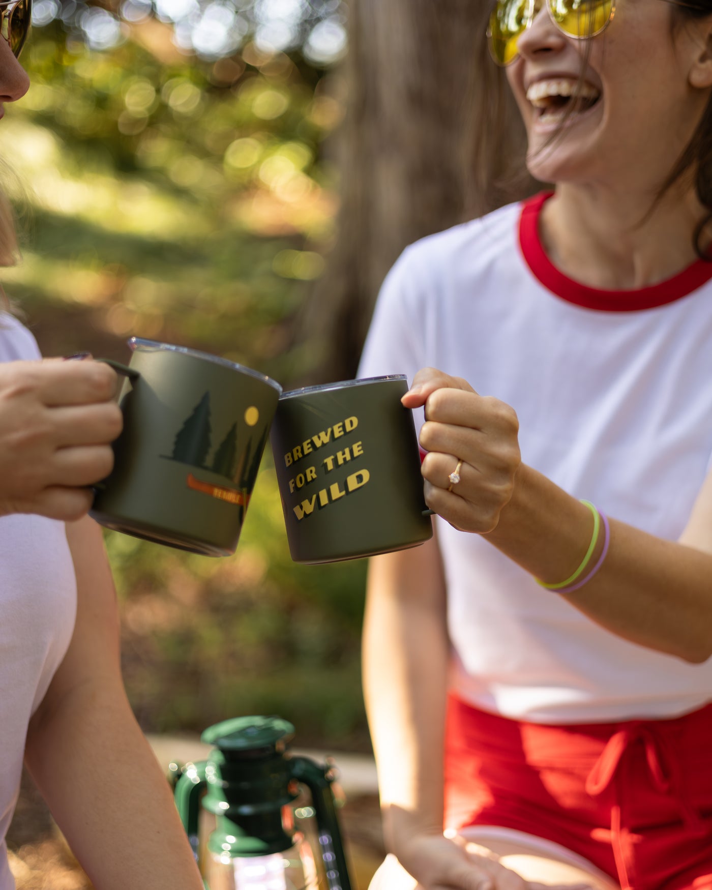 Two people outdoors, holding mugs with 'Brewed for the Wild' text, laughing.