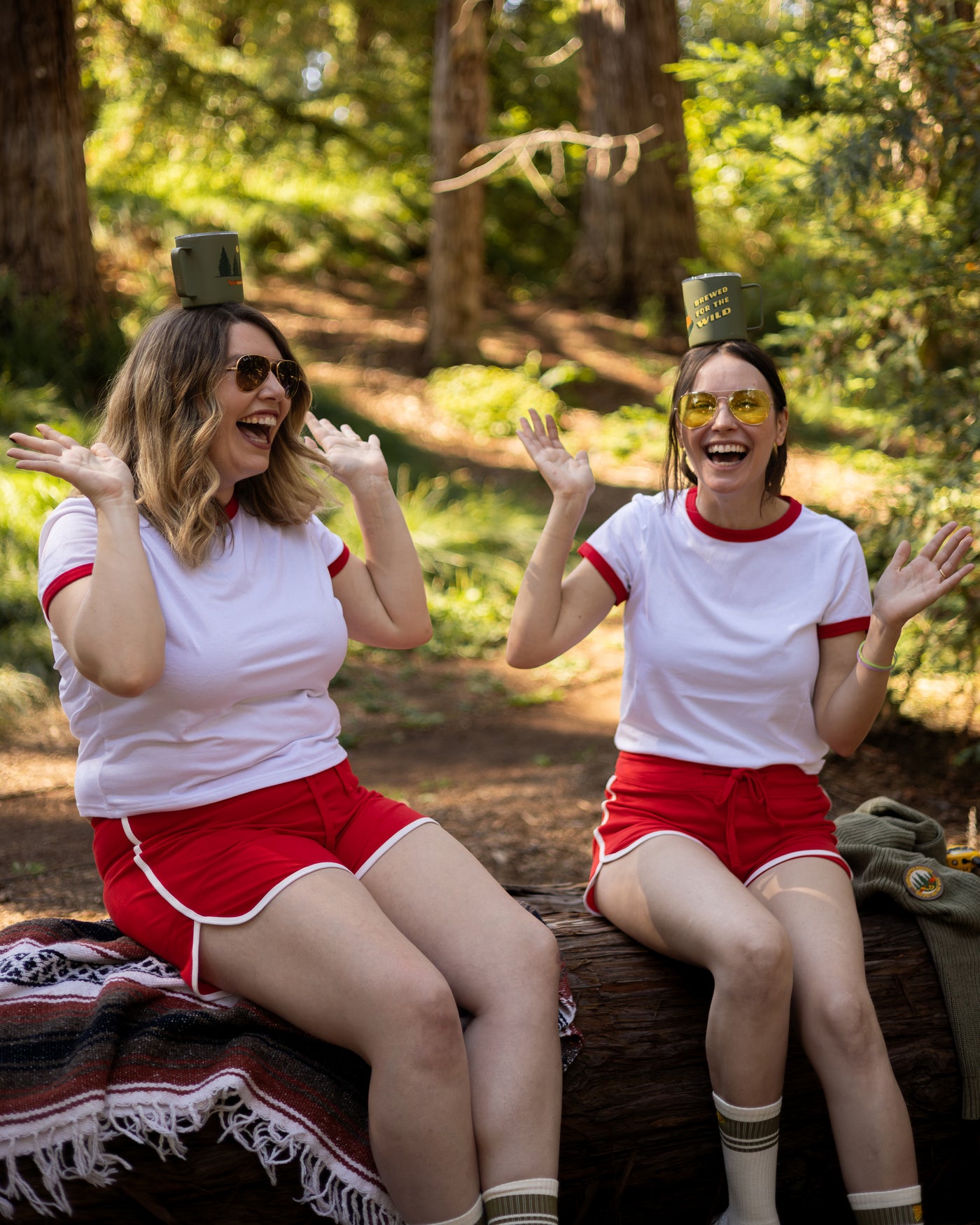 Two women in matching outfits with camp cups on their heads sitting on a log in a forest.
