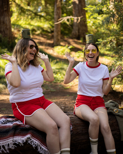 Two women in matching outfits with camp cups on their heads sitting on a log in a forest.