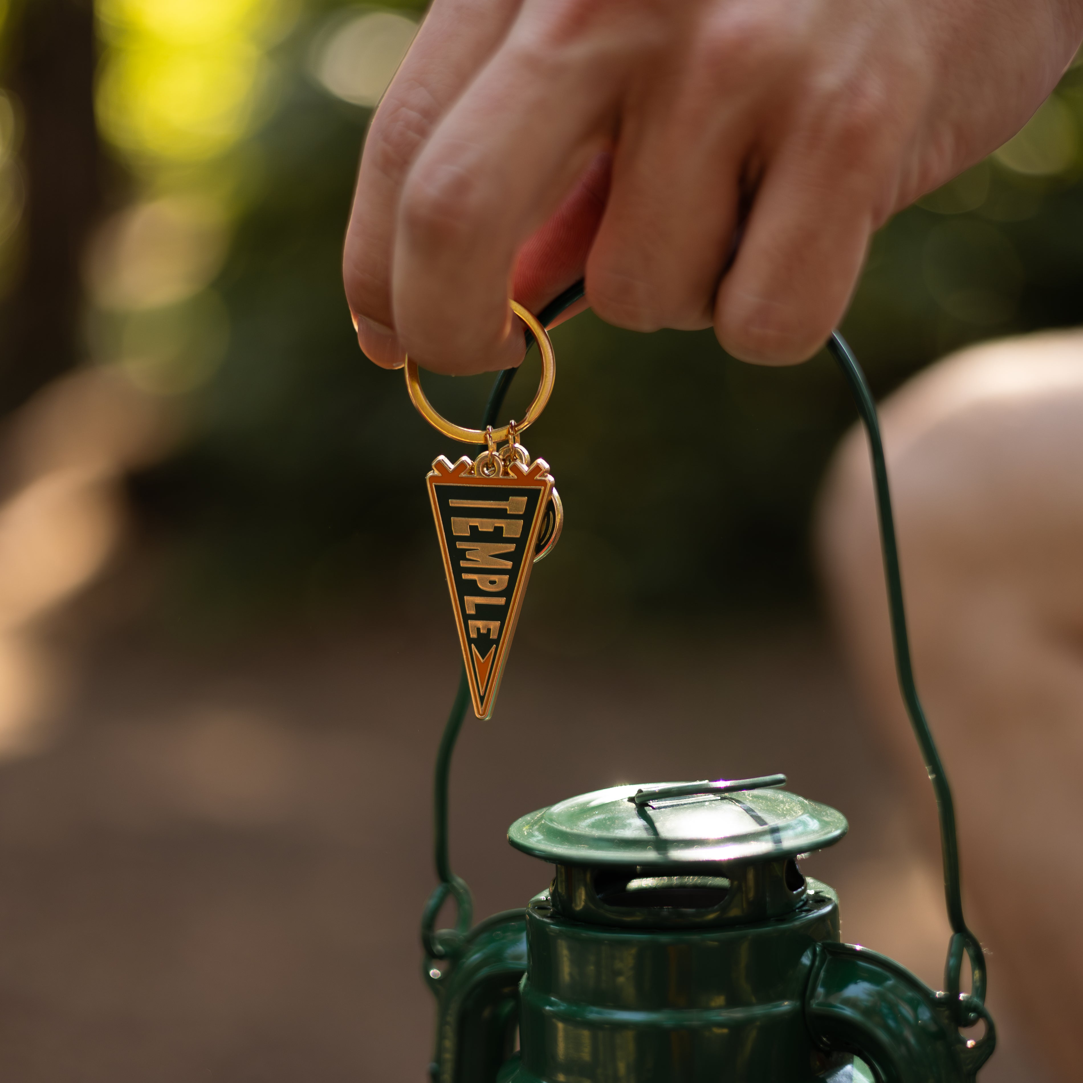 Hand holding a green lantern with a keychain featuring a pennant flag, against a blurred natural background.
