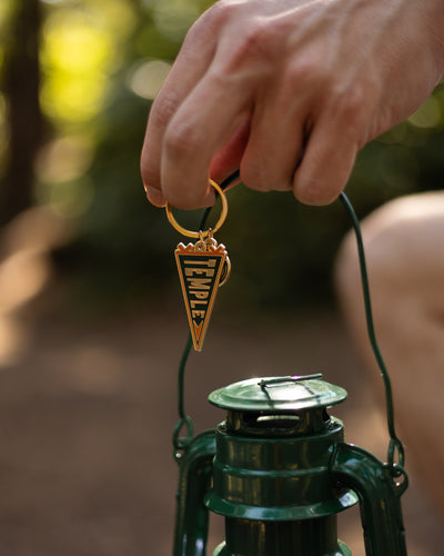 Hand holding a green lantern with a keychain featuring a pennant flag, against a blurred natural background.