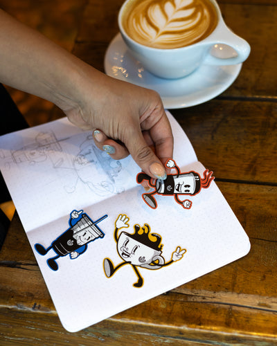 Person holding a sticker with cartoon coffee designs next to a cup of latte on a wooden table.