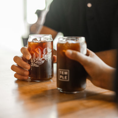 2 Peoples hands holding Glass Cans filled with cold brew coffee.