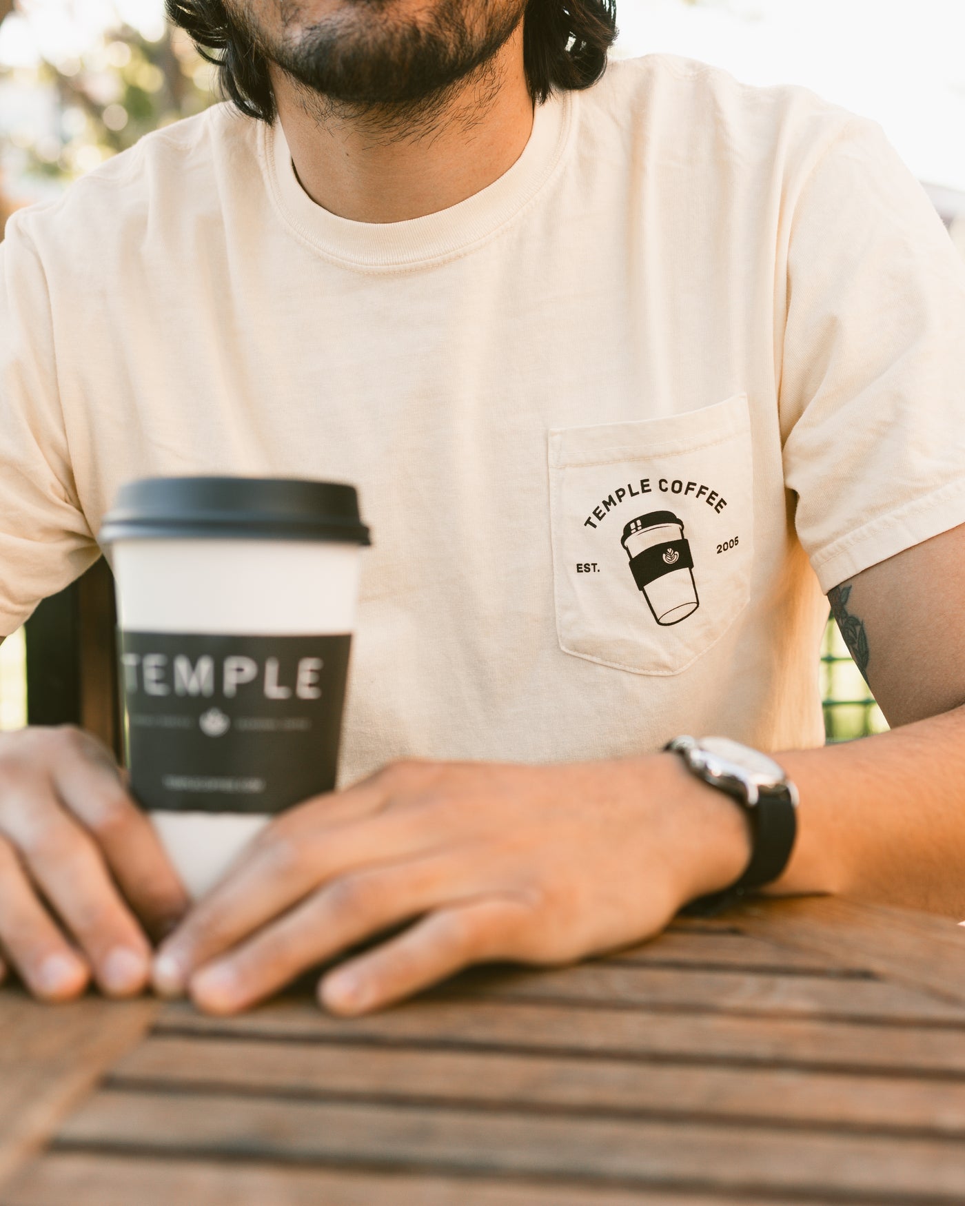 Person wearing a cream-colored T-shirt with a "Temple Coffee" logo and a to-go cup graphic on the front pocket, holding a branded Temple Coffee cup.