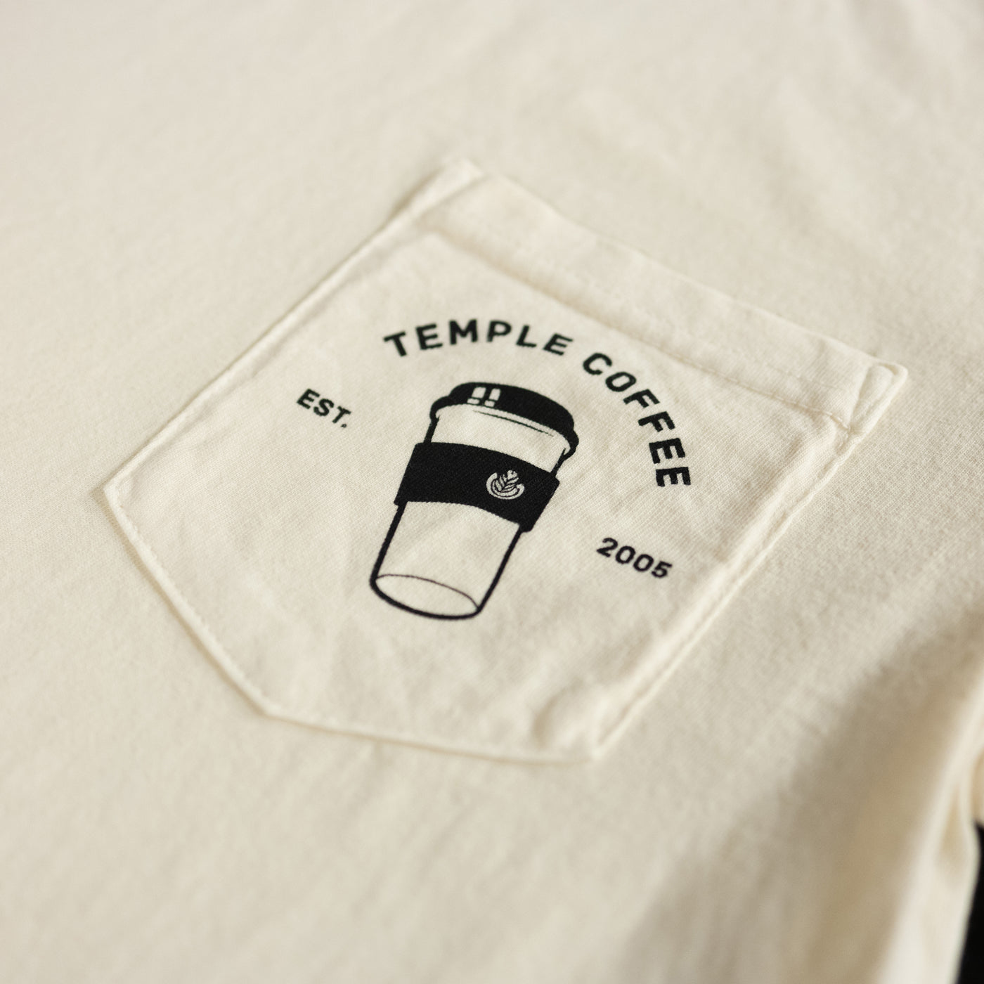 Close-up of a cream-colored T-shirt pocket featuring the 'Temple Coffee' logo with 'Est. 2005' and a to-go coffee cup graphic.