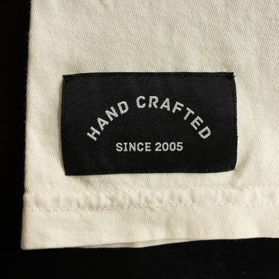 Close-up of the sewn-on 'Hand Crafted Since 2005' tag on a cream-colored T-shirt, positioned just above the hem.