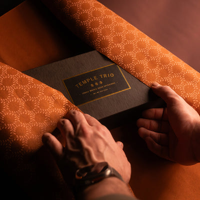 Person wrappinga black box with 'Temple Trio' branding on with orange patterned paper.