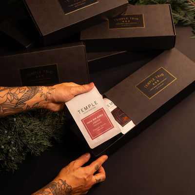 Person opening a black gift box with 'Temple' branding, surrounded by greenery.